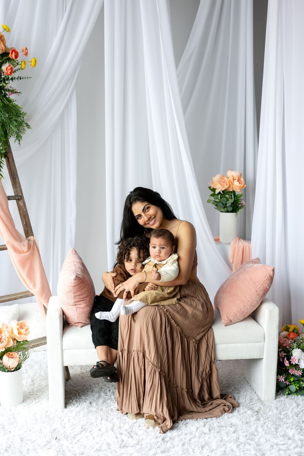 A smiling mother in a brown dress sits with her two young children in a bright floral studio.