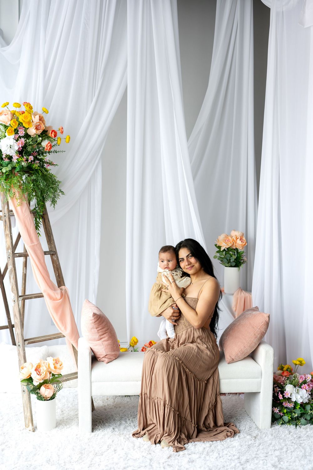 A mother in a brown dress smiles while holding her infant in a bright studio with floral decor.