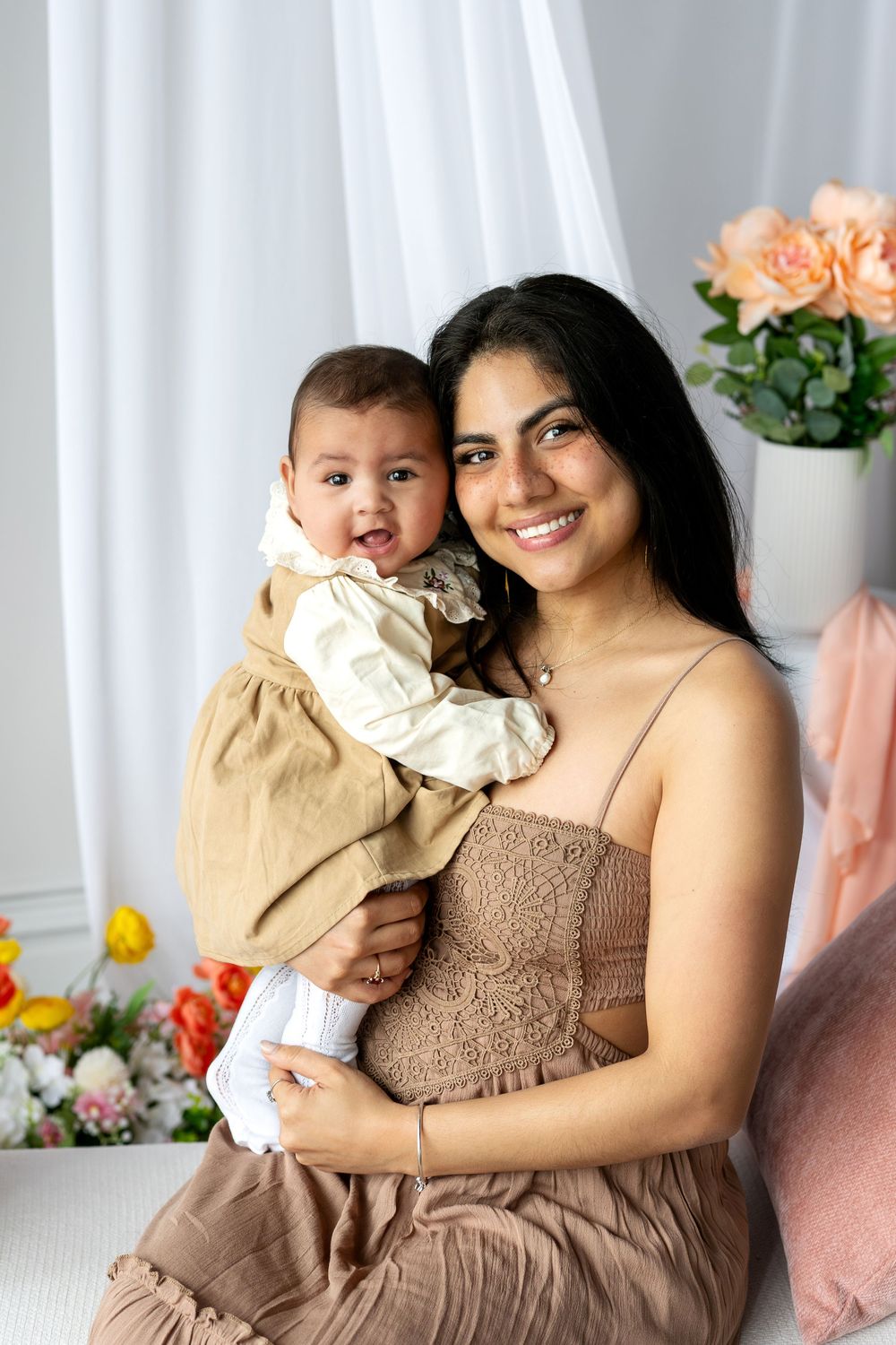 A smiling mother holds her happy baby girl in a bright studio with flowers and white curtains.