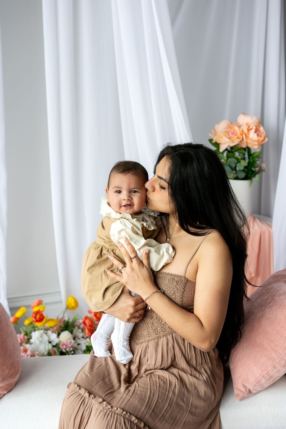 A dark-haired mother gently kisses her smiling baby in a floral studio with white draping.