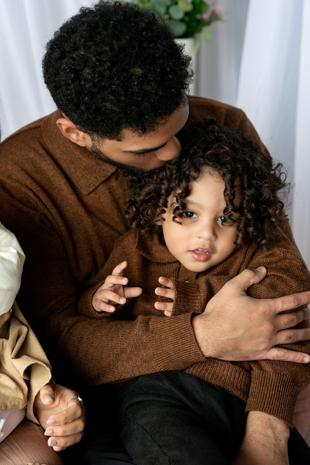 A father with curly hair tenderly embraces his young curly-haired child in a softly lit studio.