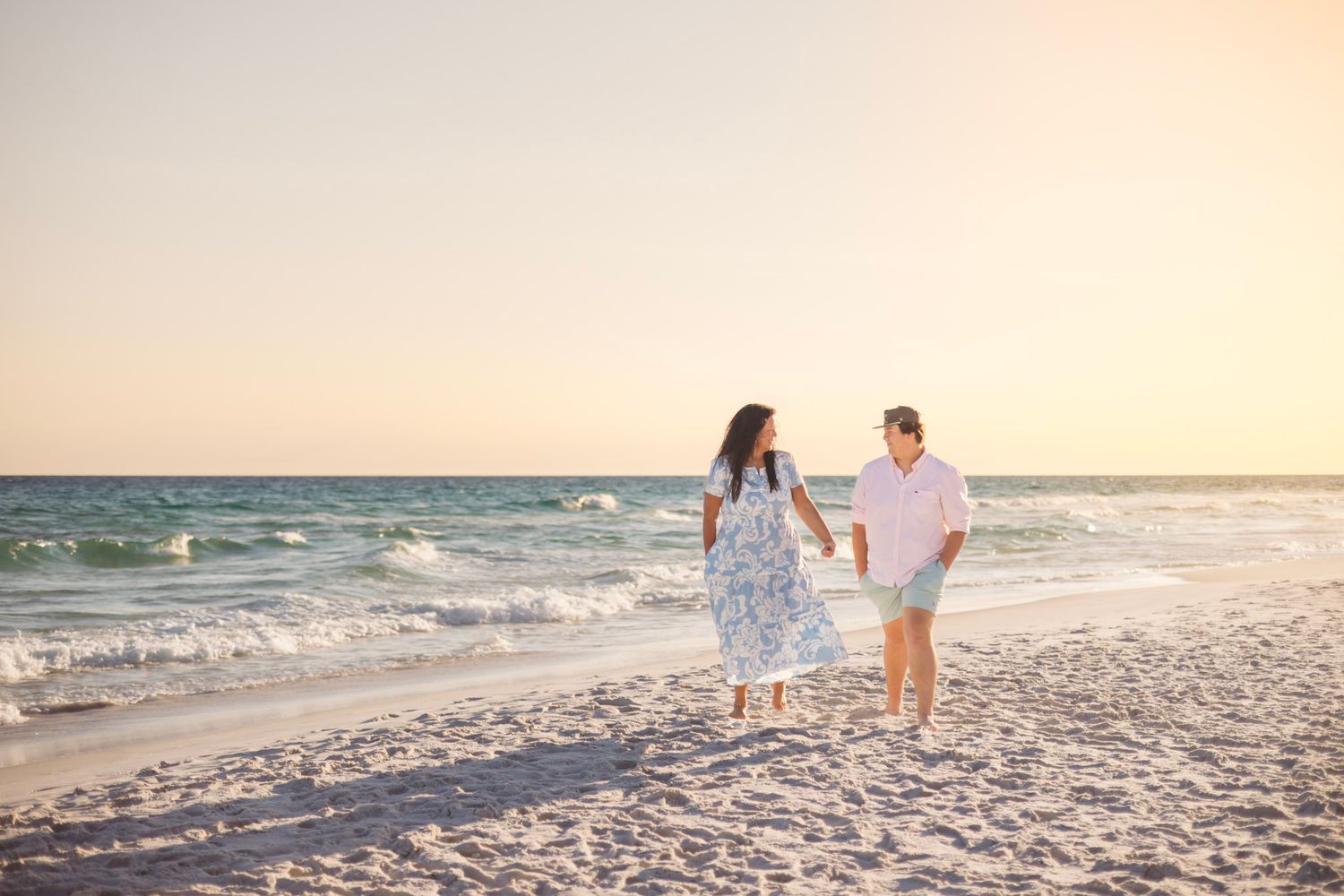Couple holds hands walking along sandy beach with turquoise water at golden sunset.