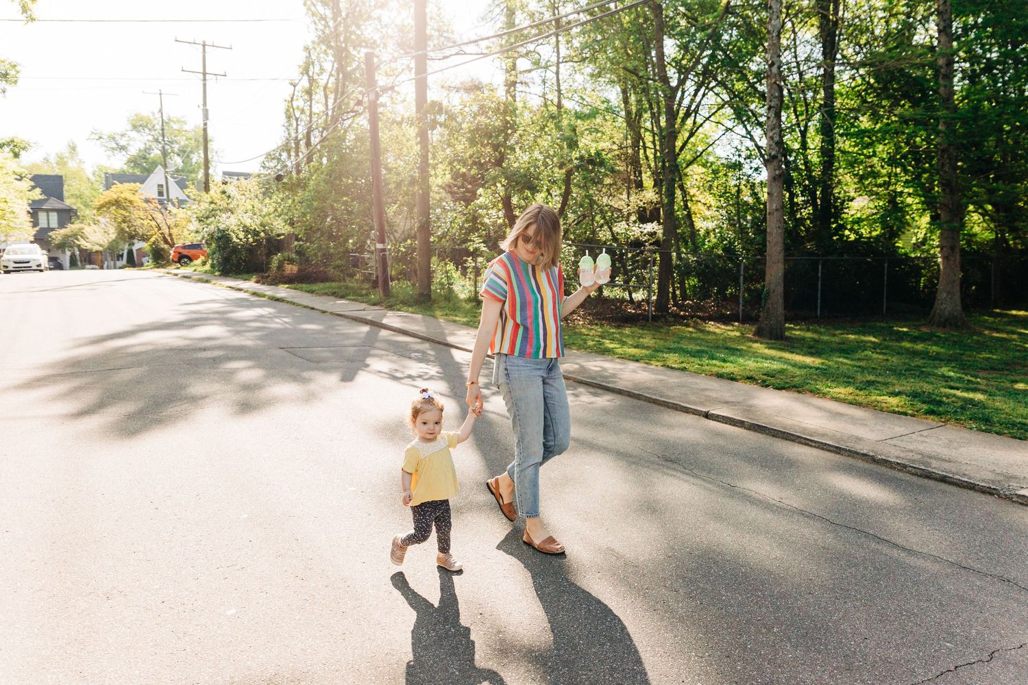 A mother and toddler holding hands while crossing a sunny suburban street lined with lush green trees in Charlotte, NC.