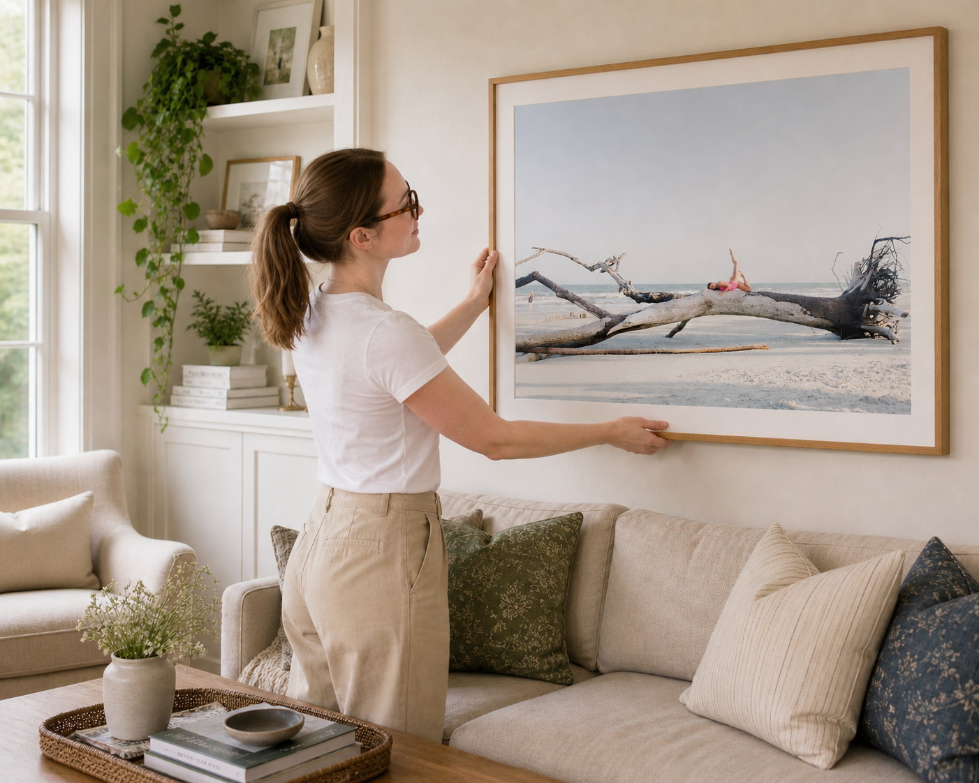 Heather Cahoon hanging a large framed coastal landscape portrait with a dancer posed in tree above a sofa in a bright modern living room.