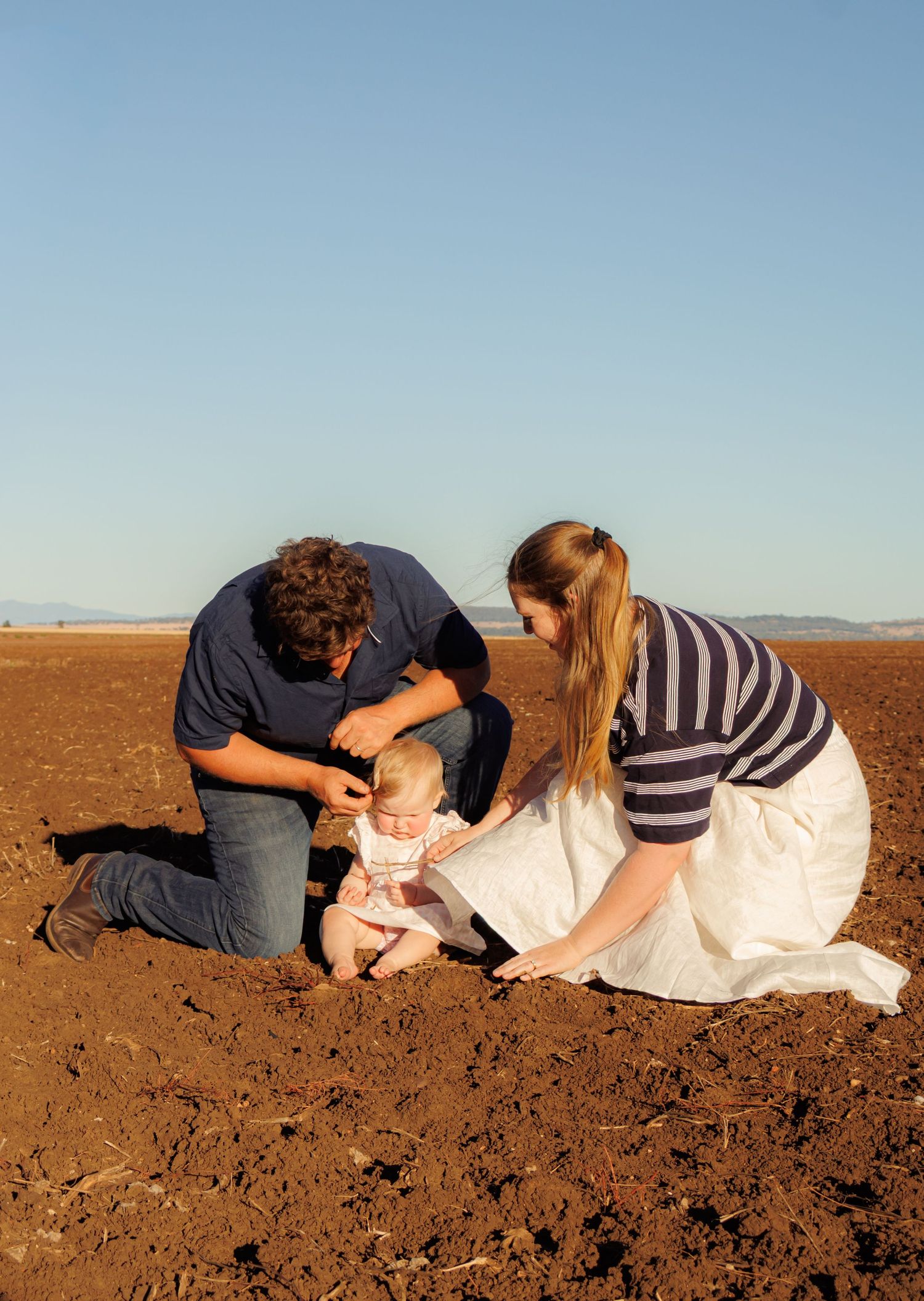 Young couple kneeling in plowed field with baby on red soil under clear blue sky.