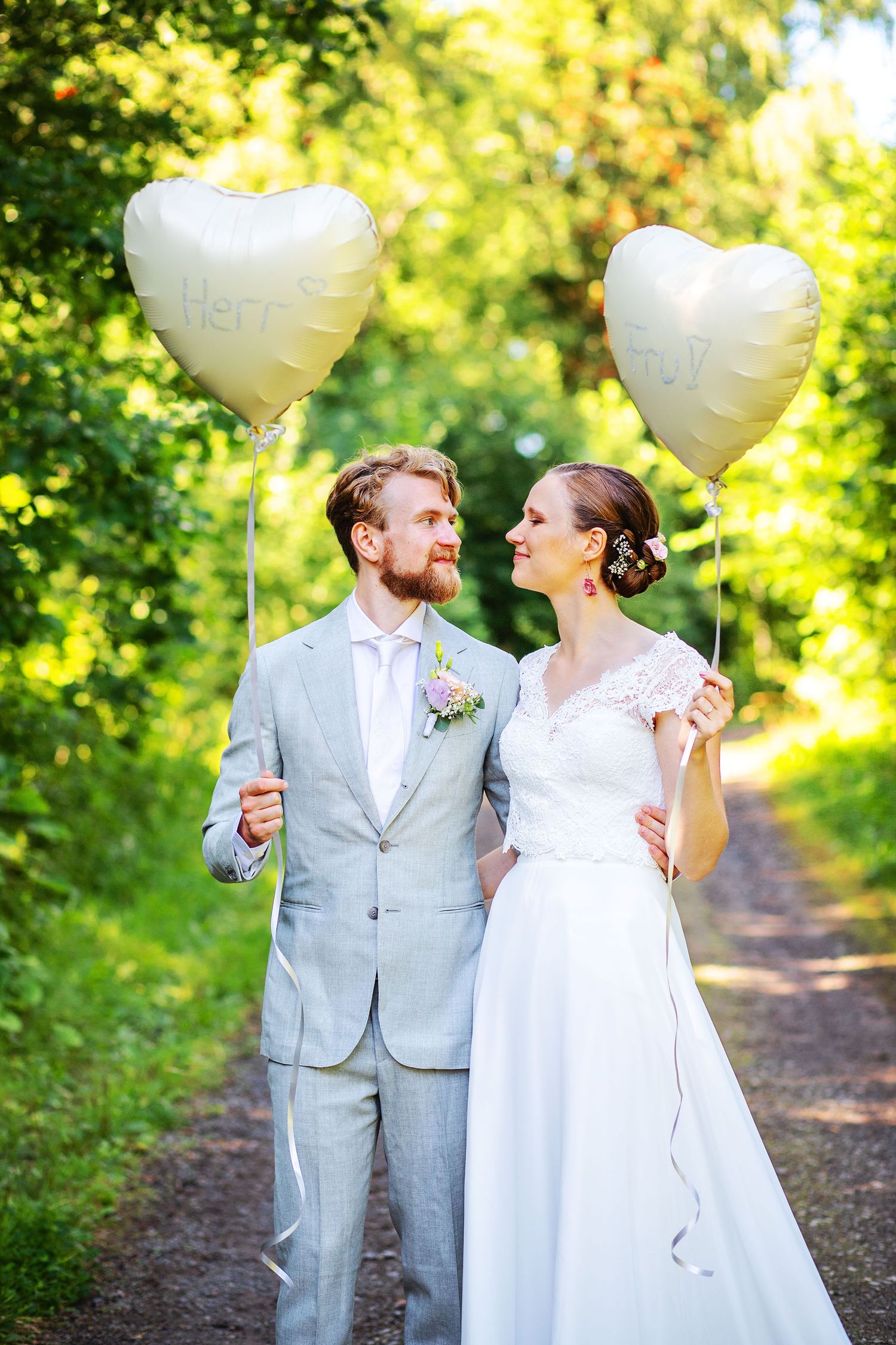 Wedding couple holding heart-shaped balloons on a tree-lined path, smiling at each other in summer greenery.