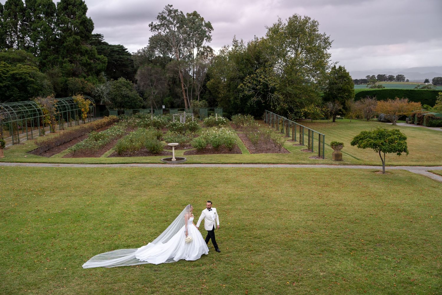 Elegant bride and groom posing separately under a garden pergola in a classic black and white wedding photo.