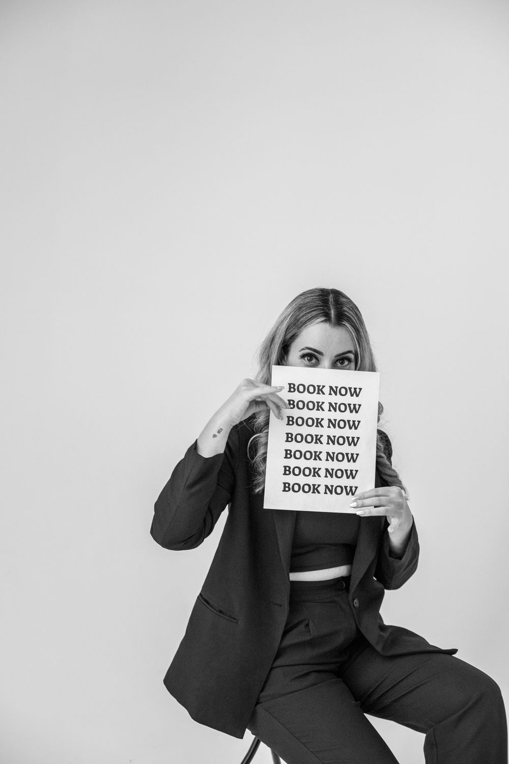 Black and white photo of a blonde woman in a suit holding a Book Now sign in front of her face while seated.