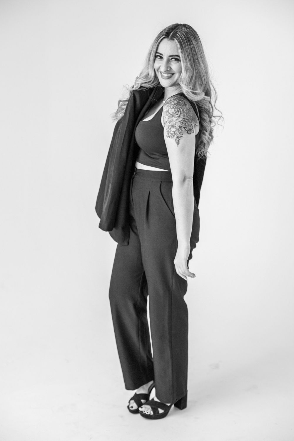 Black and white photo of a smiling blonde woman in wide-leg pants, crop top, and heels, holding a blazer over her shoulder.