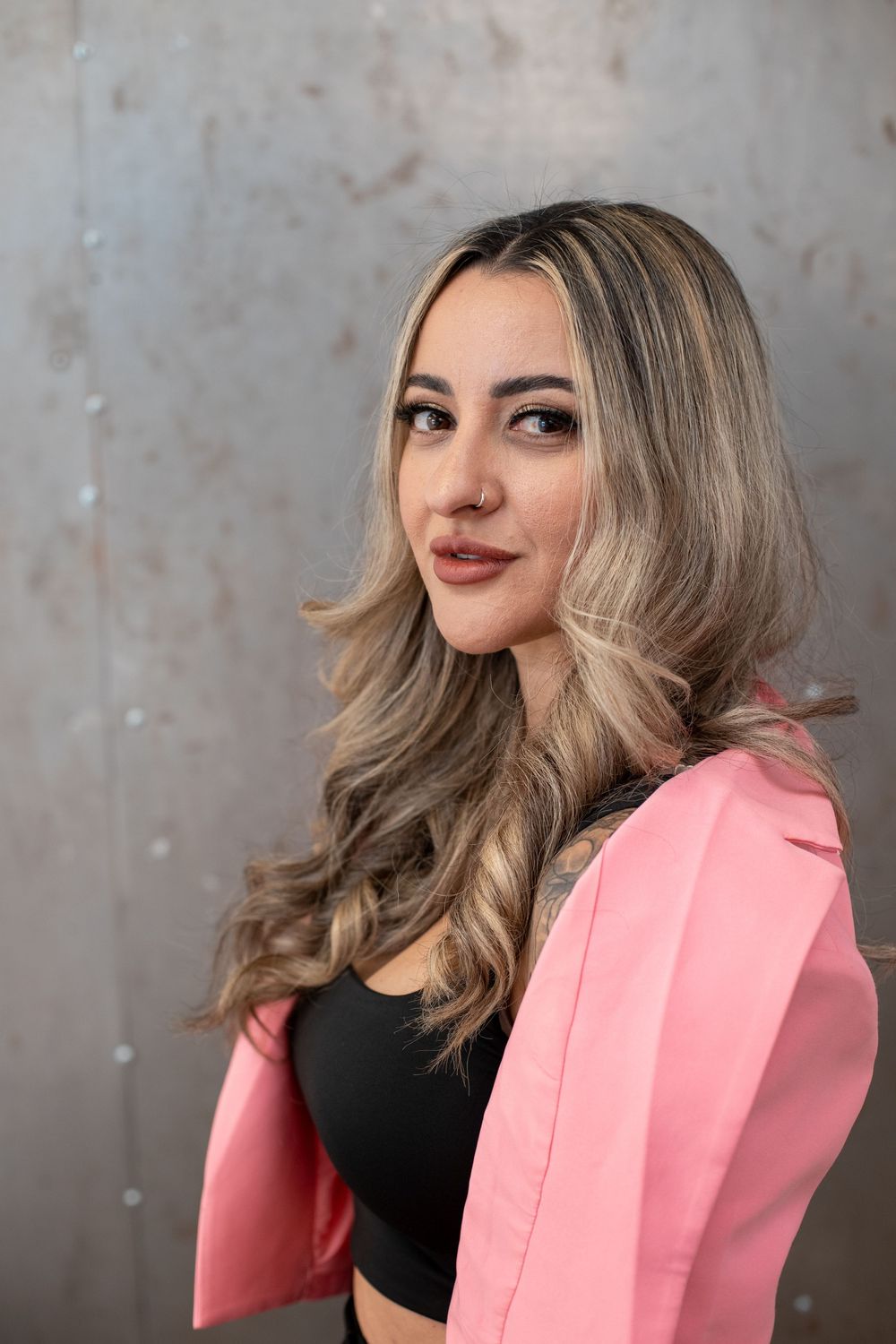 Young woman with wavy blonde hair wearing a pink blazer and black top, posing confidently against a gray wall.