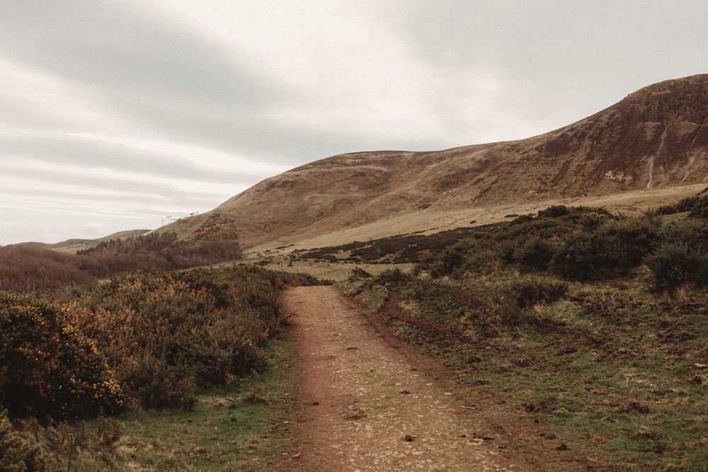 A winding dirt trail through brown heather moorland leading toward rolling hills under an overcast sky.
