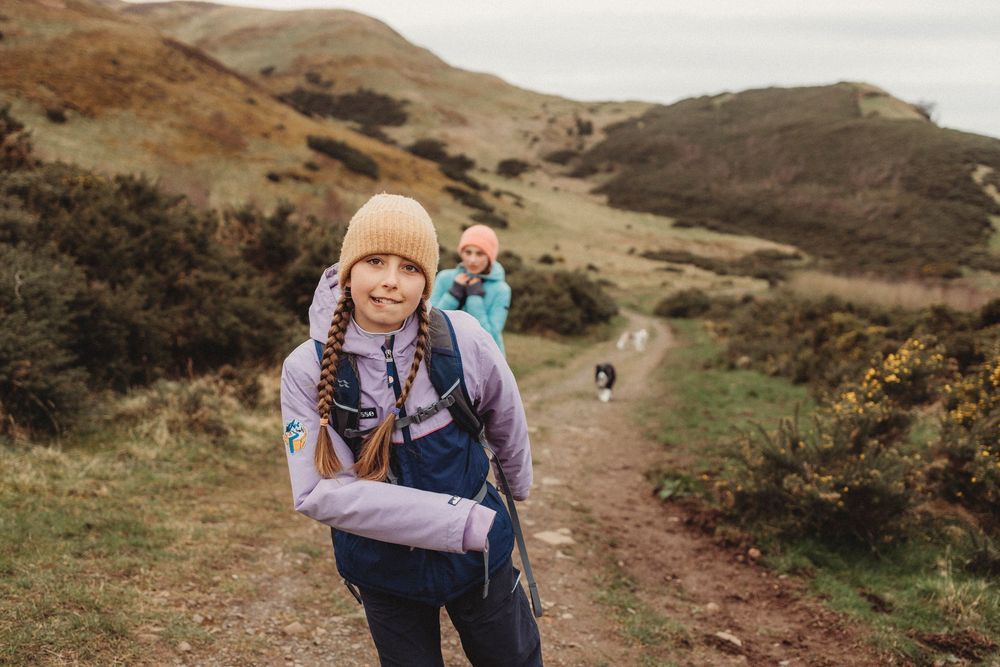 Two hikers with backpacks walking along a scenic mountain trail surrounded by golden hills and lush greenery.
