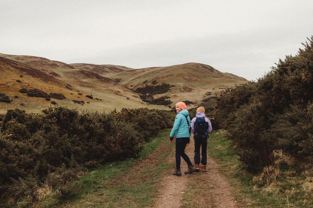 Two hikers walking along a scenic trail through rolling hills and lush greenery on a cloudy day.