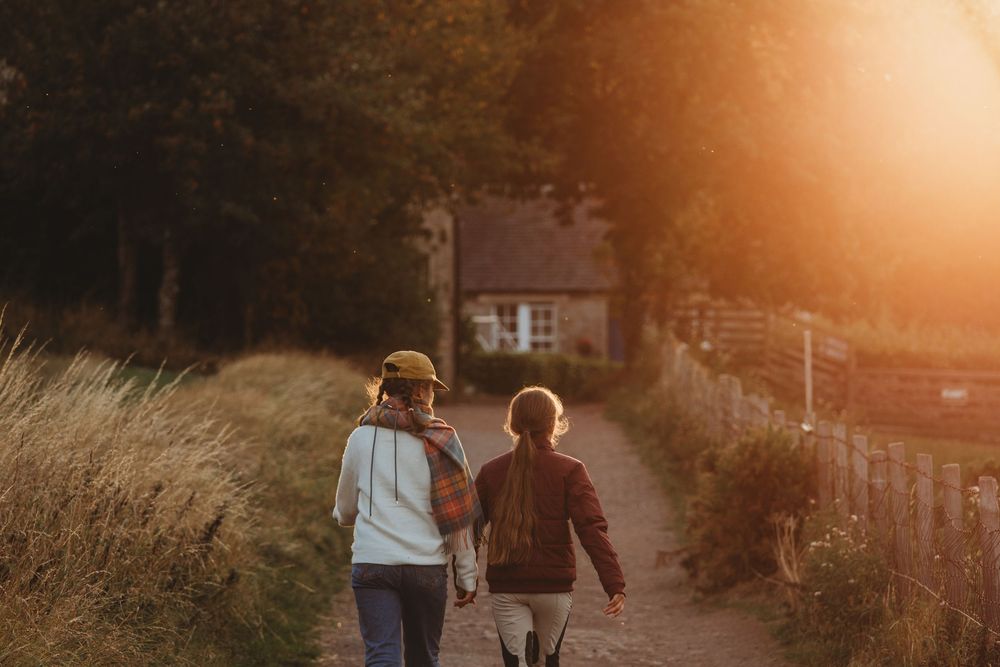 Two children walk hand-in-hand down a sunlit path toward a cottage, surrounded by golden autumn trees.