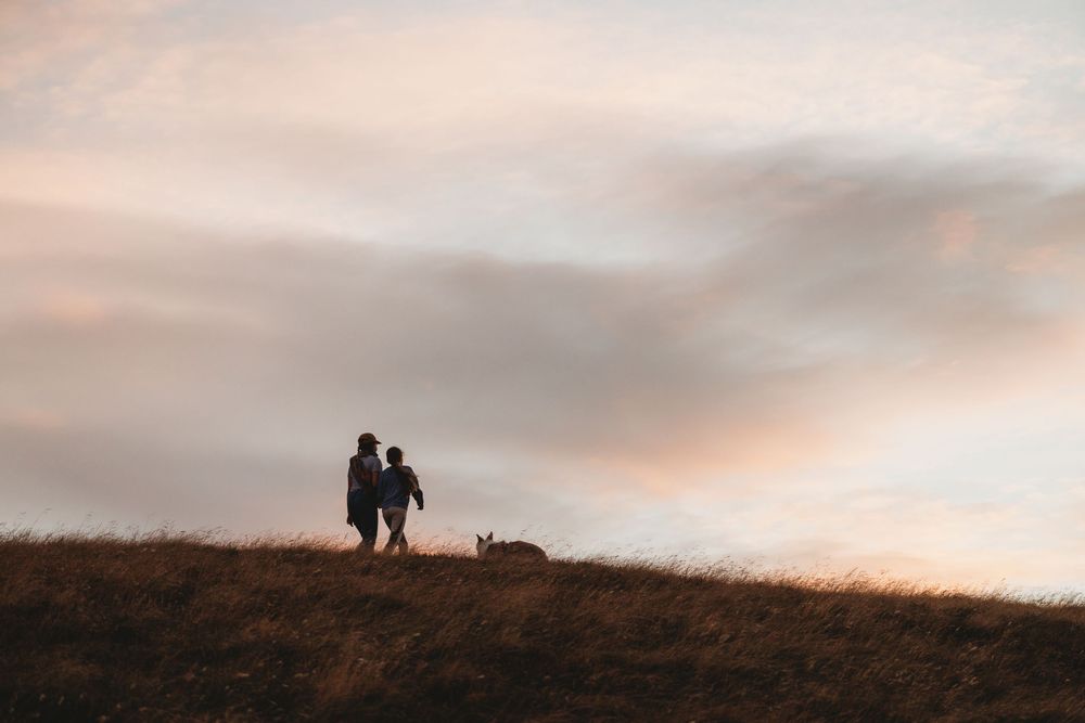 Silhouette of a couple and their dog standing on a hill at sunset with a dramatic pink and blue sky.