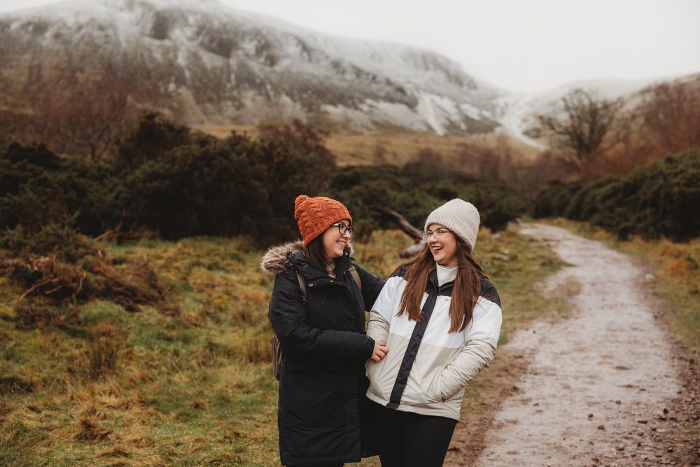 Two women in winter hats laugh together on a misty mountain trail in autumn.