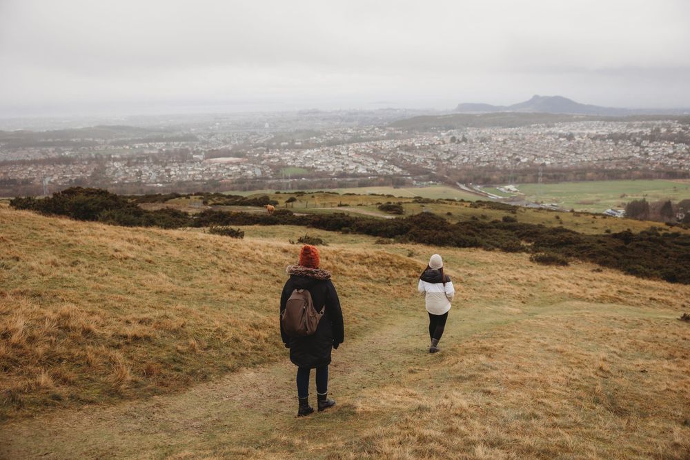 Two hikers walk along a grassy hillside trail overlooking a misty city landscape on a cloudy day.
