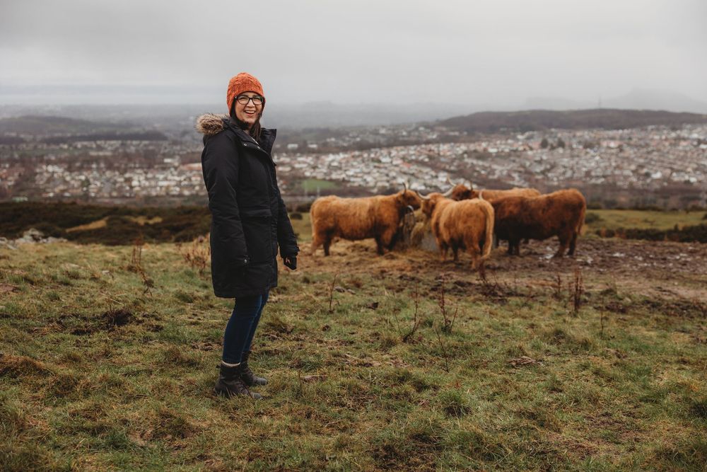 Woman in orange beanie stands near Highland cattle on a hill overlooking a city.