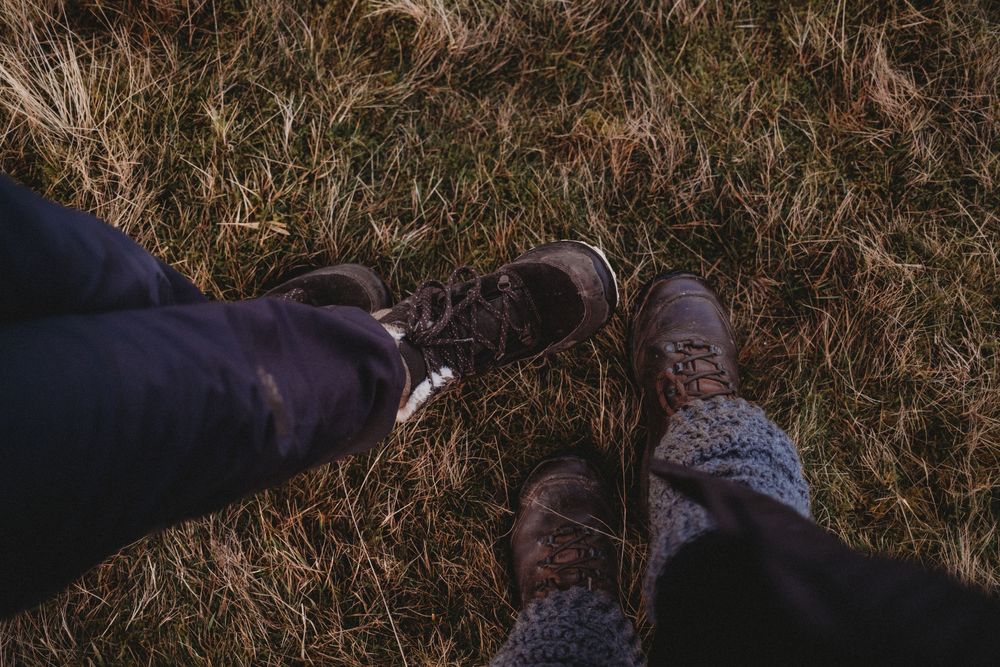 Two people sitting on dry grass wearing brown hiking boots and dark pants, viewed from above in a moody outdoor setting.