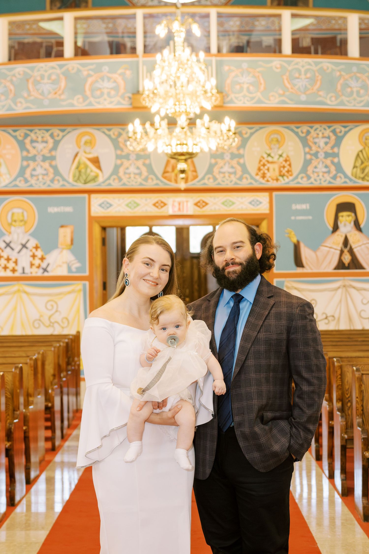 A couple in formal attire holds their baby in white christening gown inside an Orthodox church with iconic murals.