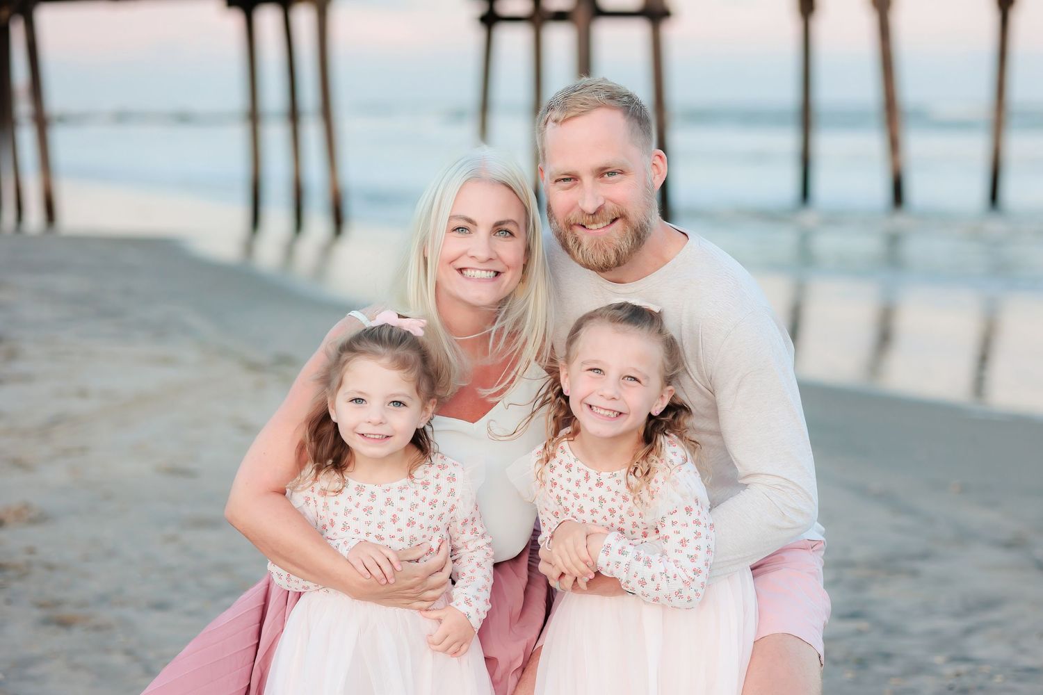 Family of four smiling together on a beach near a pier at sunset, parents holding two young girls in matching pink dresses.