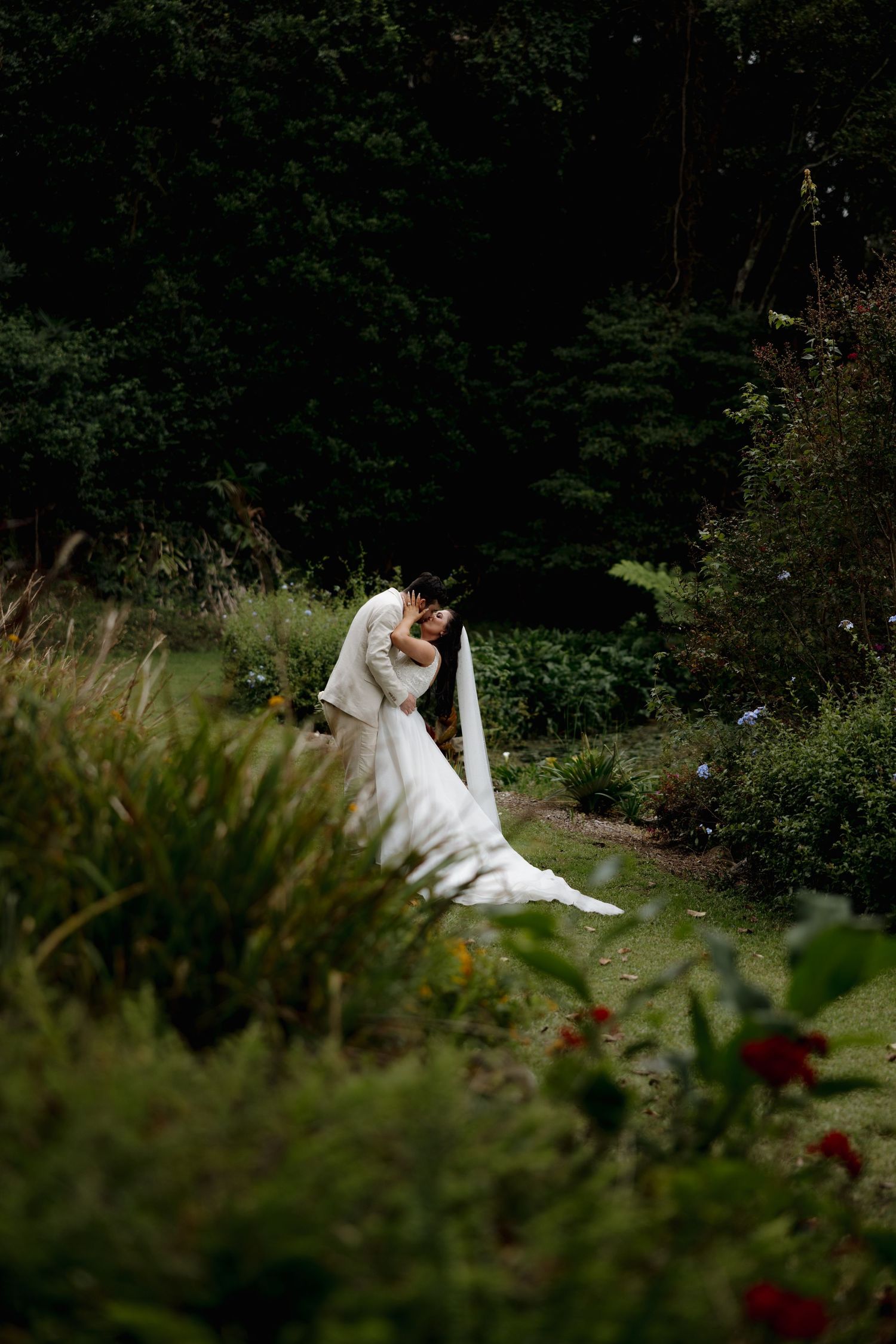 Bride and groom share a romantic kiss in a lush garden, with her white gown and veil trailing on the ground.