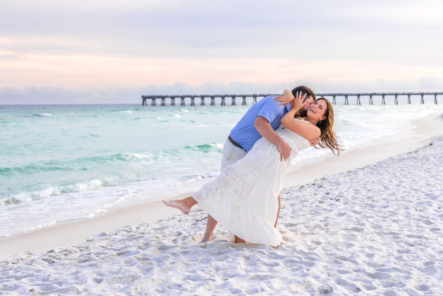 Couple sharing a romantic dip kiss on a turquoise beach at sunset, man in blue shirt, woman in white dress.