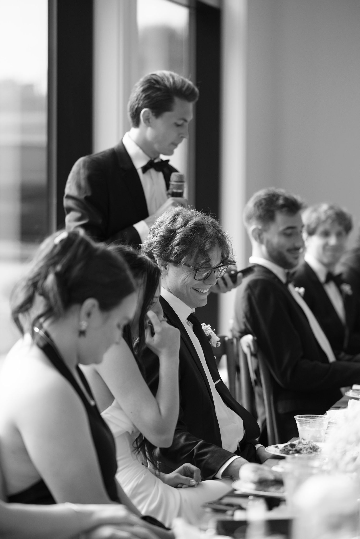 Wedding party laughs during heartfelt speeches at elegant black and white reception dinner.