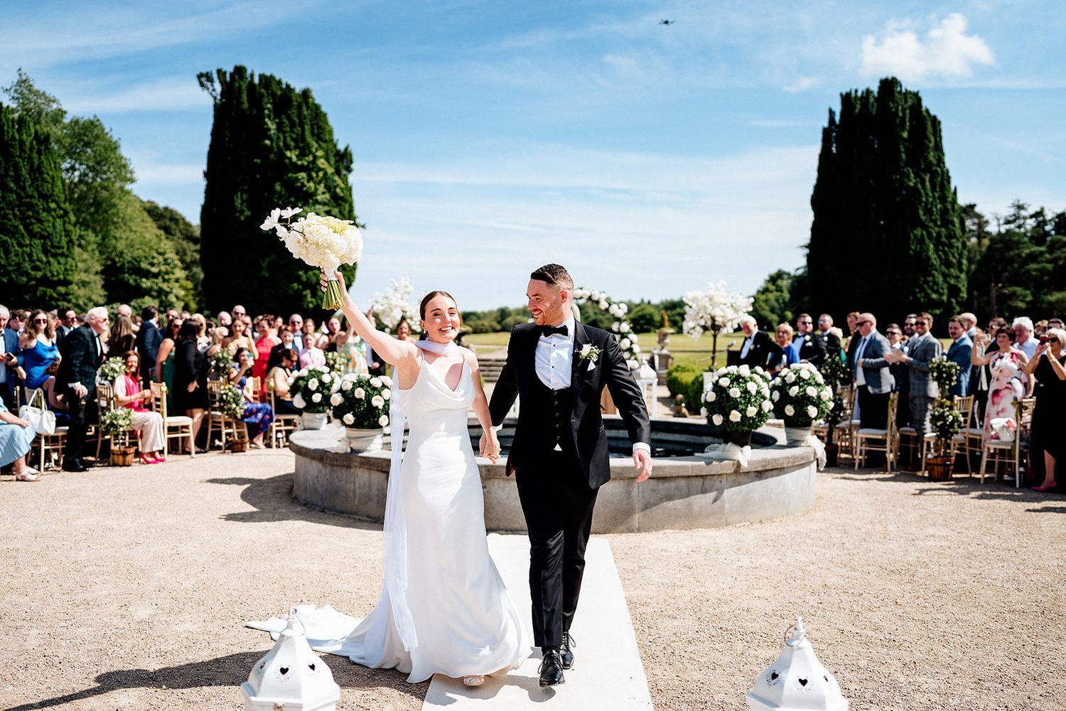 A panoramic view of an outdoor wedding ceremony with guests seated on white chairs in a garden setting under blue skies.