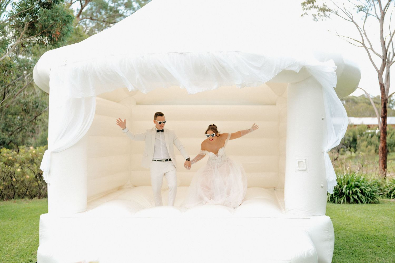 Bride and groom joyfully jumping in a white inflatable bounce house at an outdoor wedding reception.