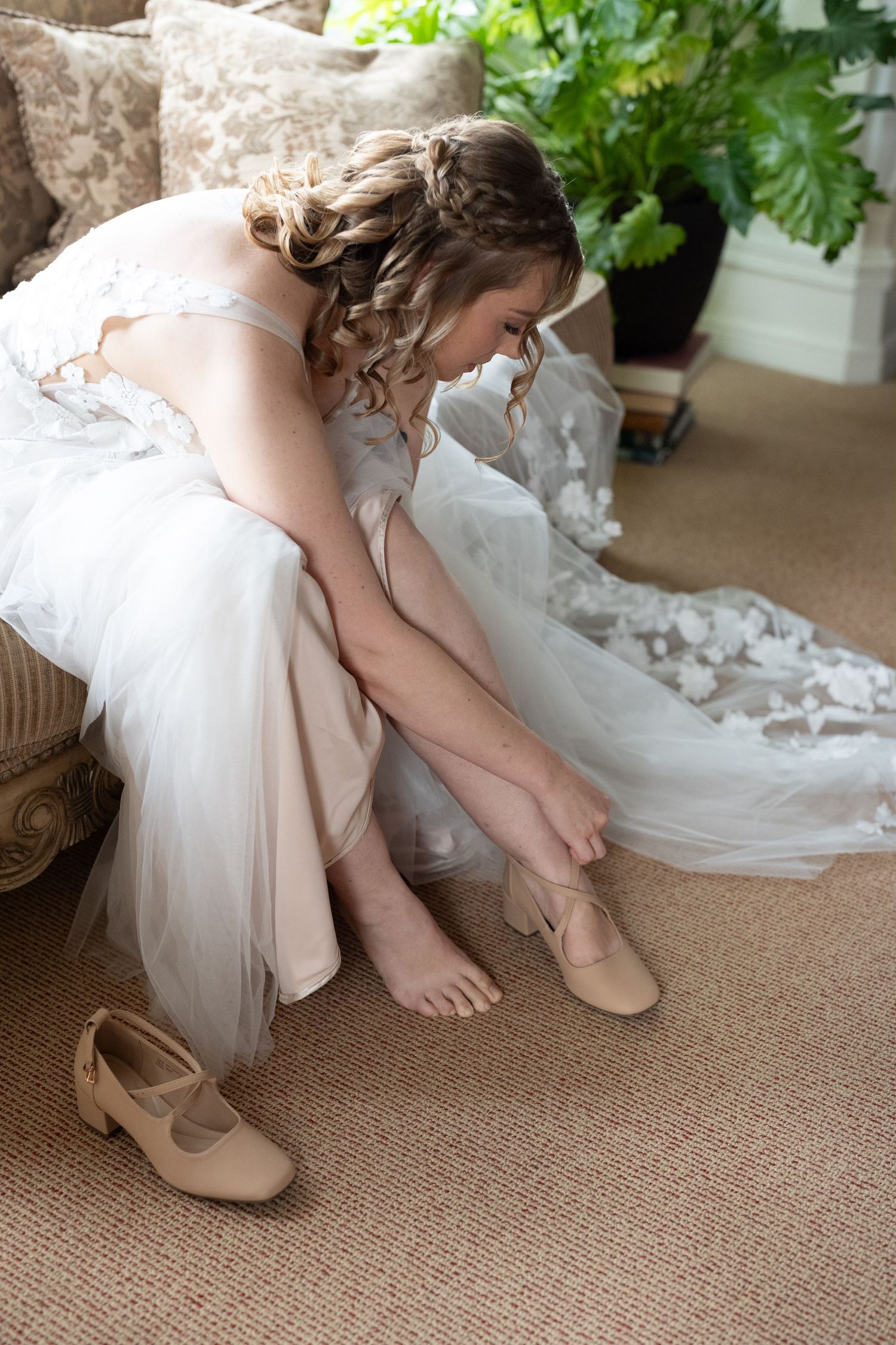Bride in white lace gown sitting on ornate chair putting on nude heels while getting ready for wedding.