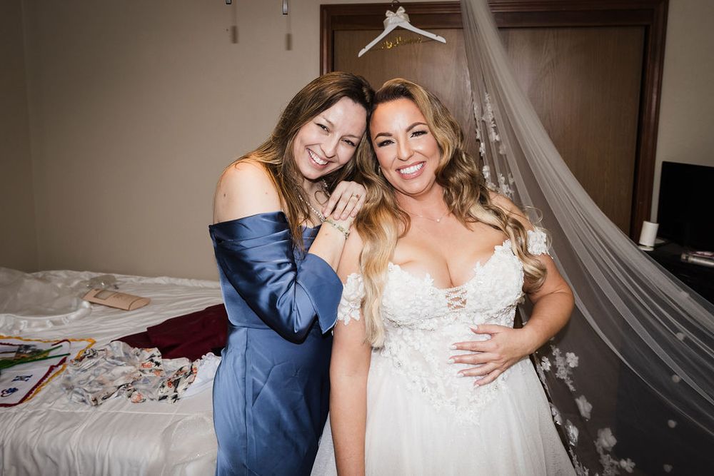 Bride in white lace gown and veil hugs smiling bridesmaid in blue dress in hotel room before wedding ceremony.