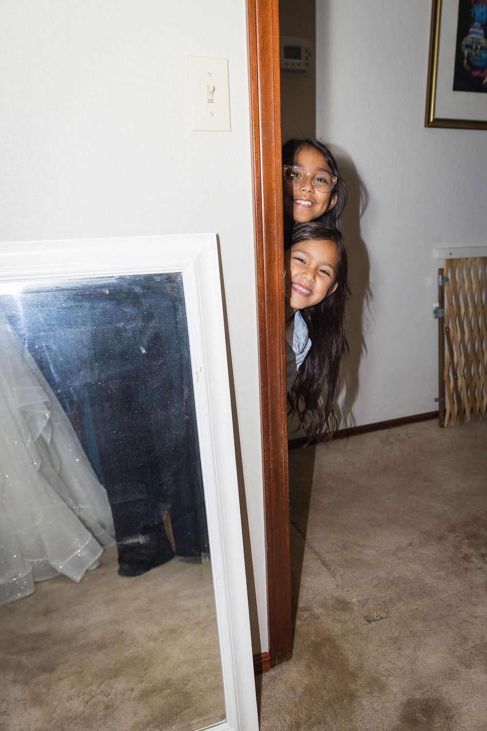 Two smiling girls peek around a wooden door frame inside a home with carpeted floors.