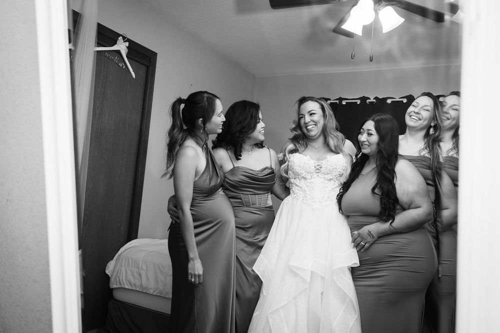 Black and white photo of a bride in a white gown laughing with her bridesmaids in matching dresses.
