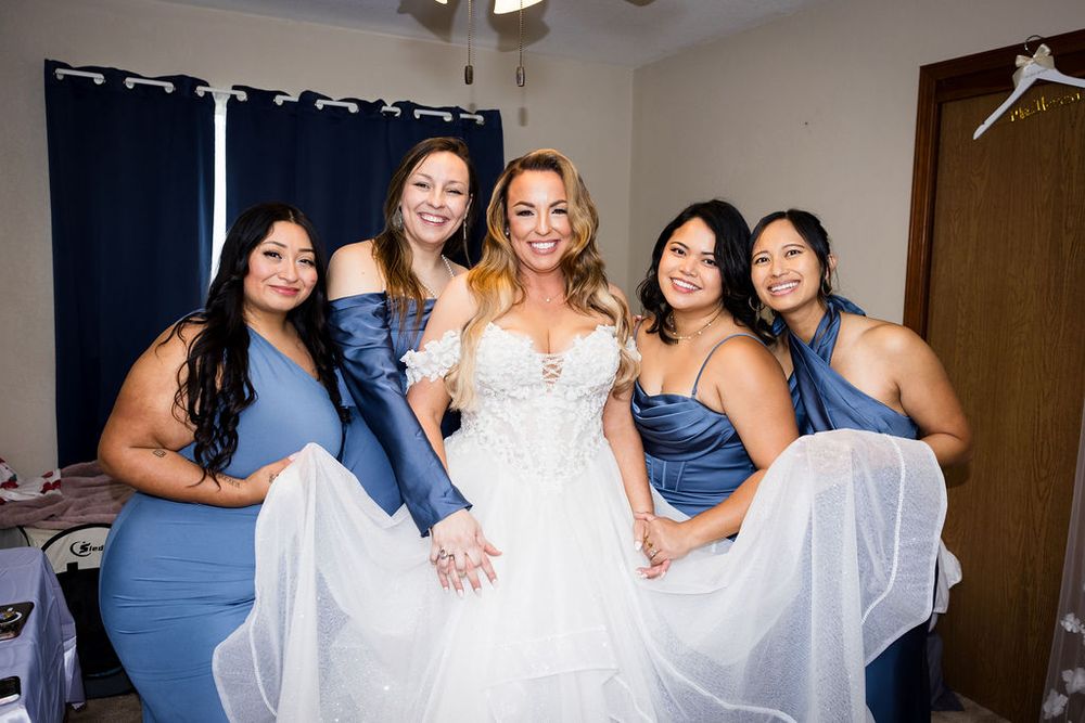 Bride in white lace gown poses with four bridesmaids in blue dresses lifting her skirt indoors.