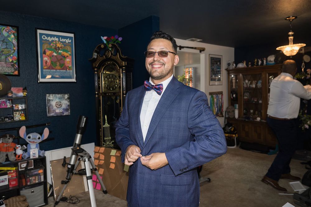 Smiling groom in navy blue suit and bow tie buttoning jacket in decorated room before wedding.