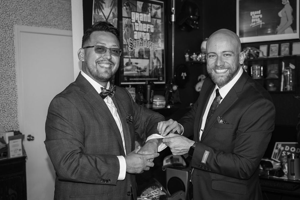 Black and white photo of two grooms in suits smiling and exchanging rings indoors.