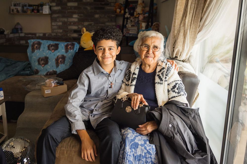 Young boy and elderly woman smiling together on a couch by a window.