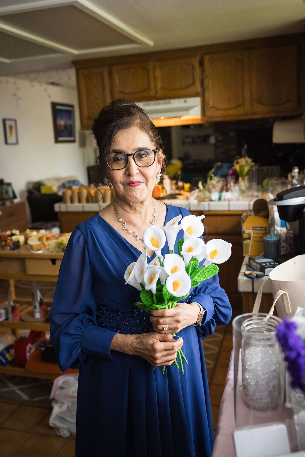 Elegant older woman in blue dress holding white calla lily bouquet in kitchen.