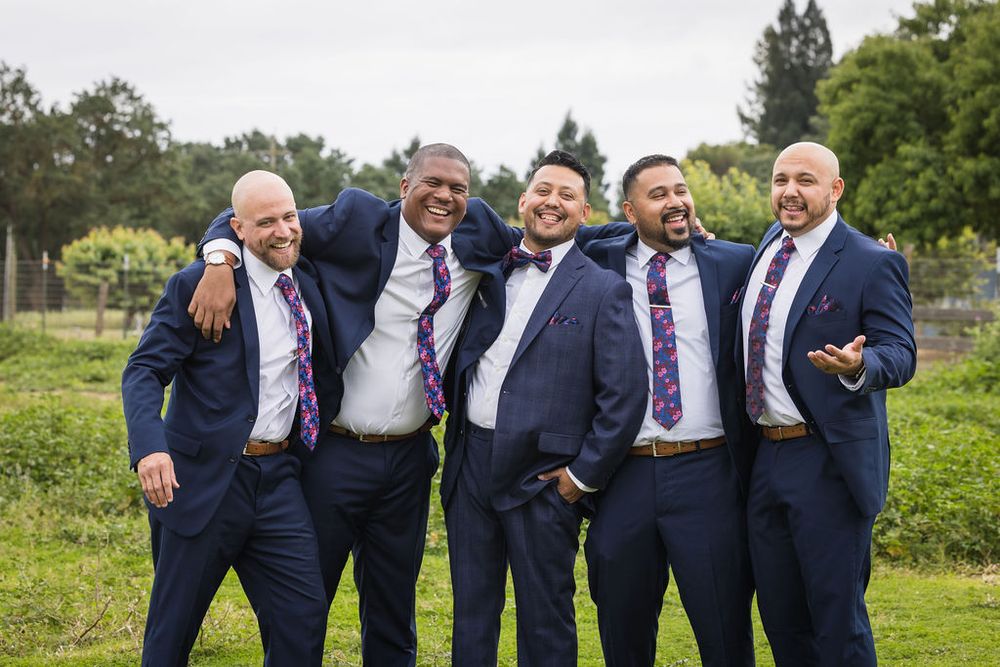 Five men in matching navy blue suits and purple ties pose together outdoors at a wedding.