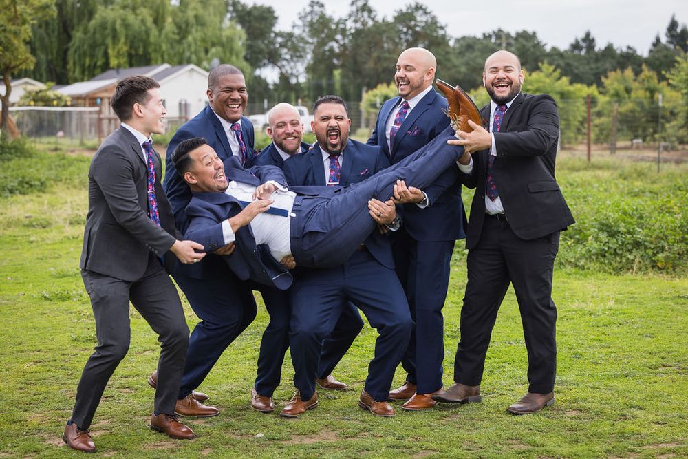 Groomsmen in navy suits playfully lifting the groom outdoors on a green lawn.