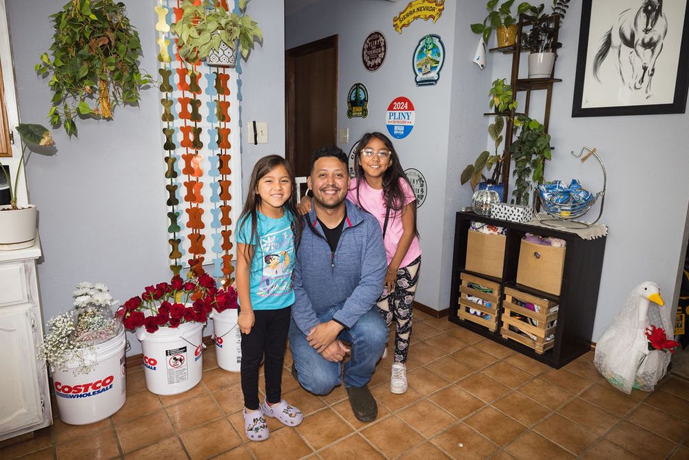 Man kneeling with two young girls smiling inside home entryway decorated with plants and colorful ornaments.