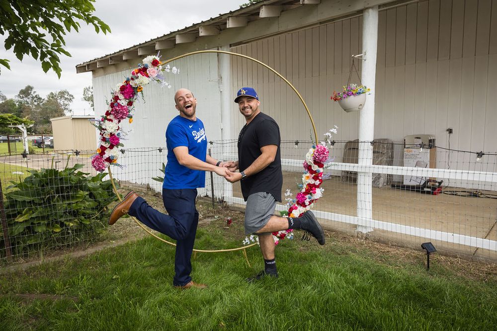 Two men playfully pose with floral hoop wedding decoration on green lawn outside barn venue.
