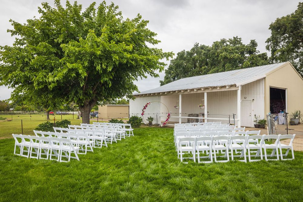 Outdoor wedding ceremony setup with white chairs on green lawn beside rustic barn venue.