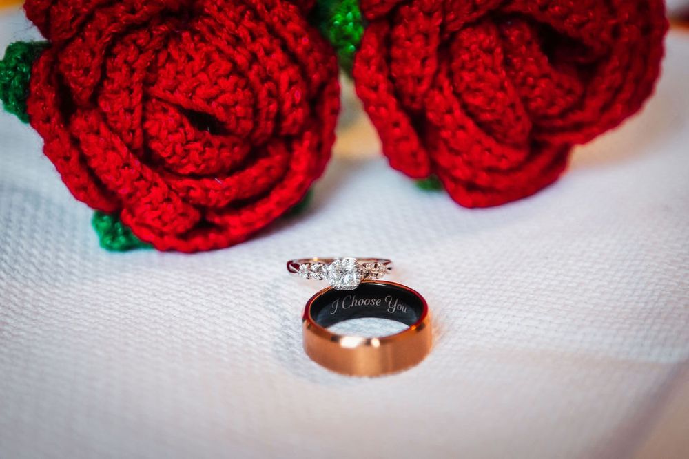 Two gold wedding rings beside red crocheted roses on white fabric, one engraved with I Choose You.