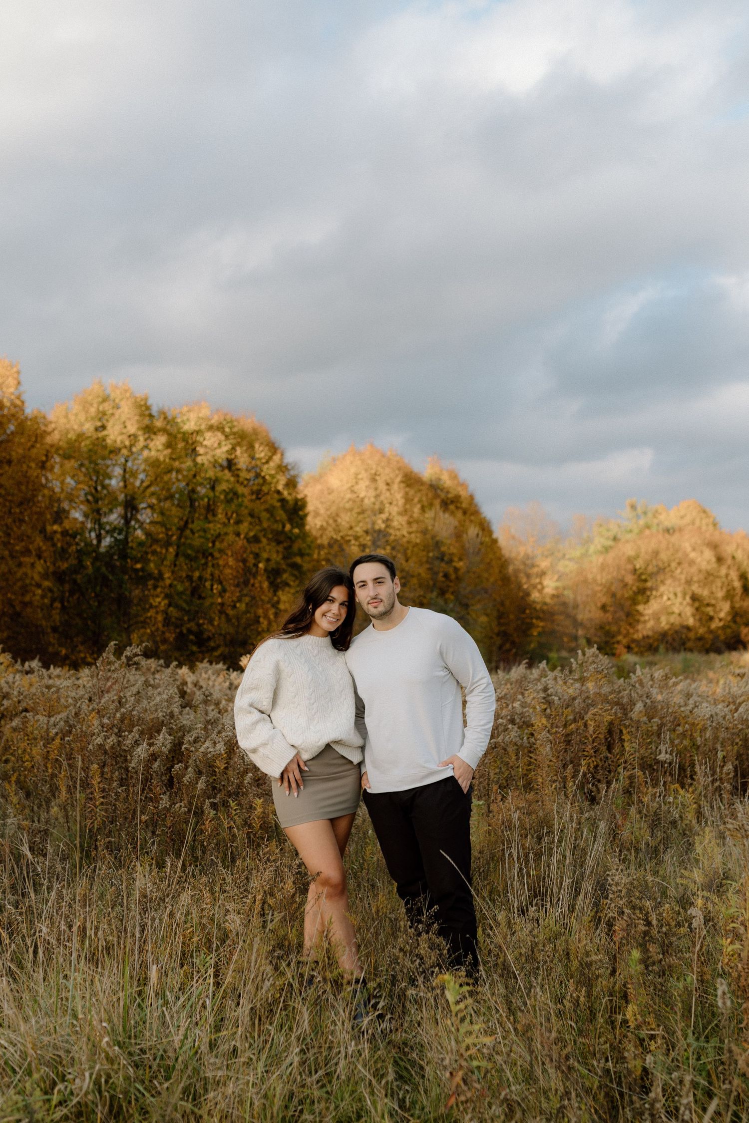 Couple in white sweaters posing in an autumn field with golden trees and cloudy sky backdrop.