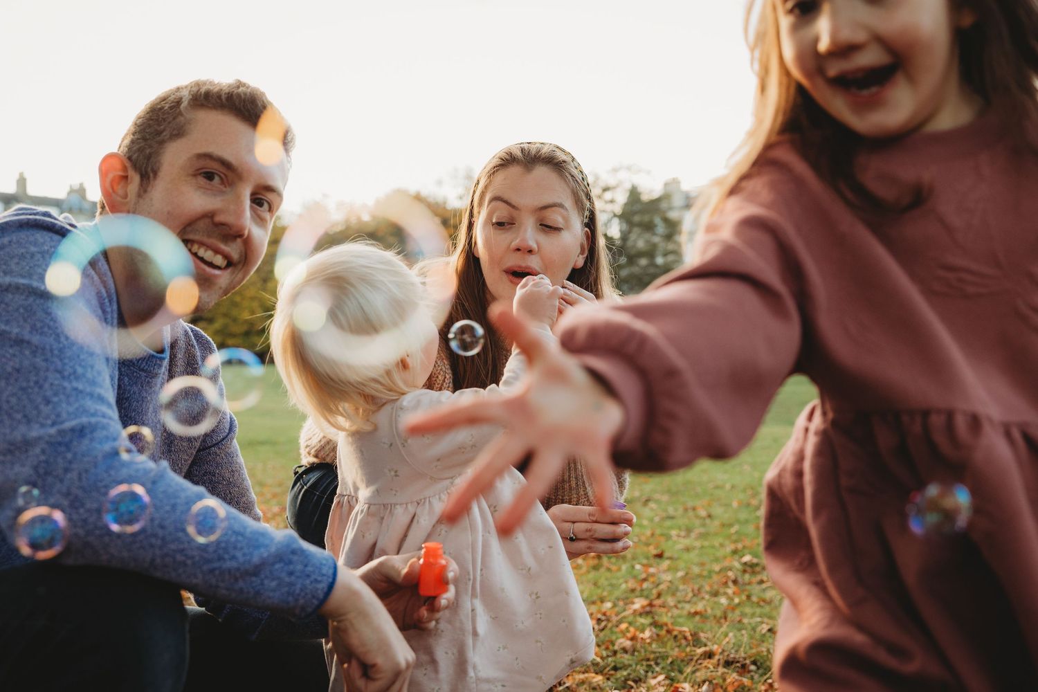 Family playing with bubbles outdoors in a park, parents and two young girls laughing and reaching for floating bubbles.