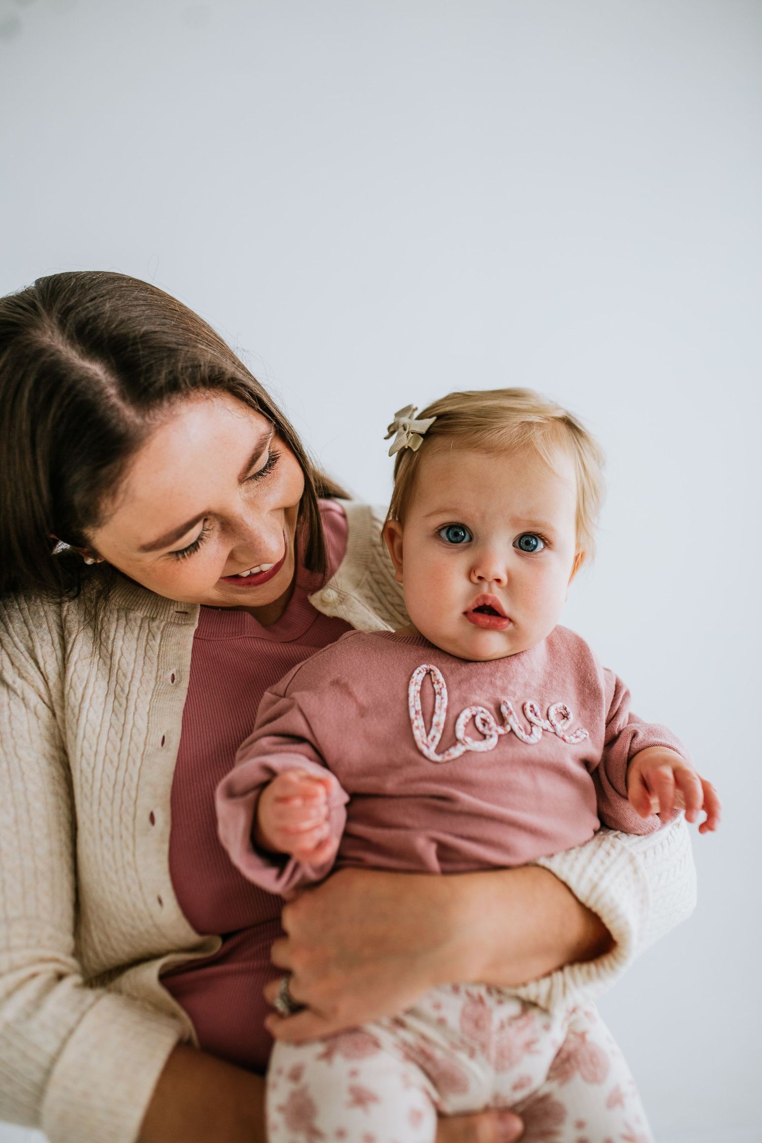 A mother in a cream sweater holds her baby girl wearing a pink love sweatshirt and floral pants.