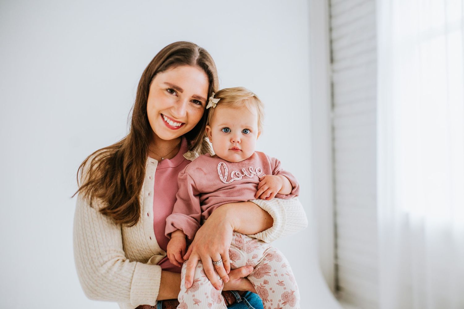 A brunette mother smiling while holding her baby girl in a pink love sweatshirt.