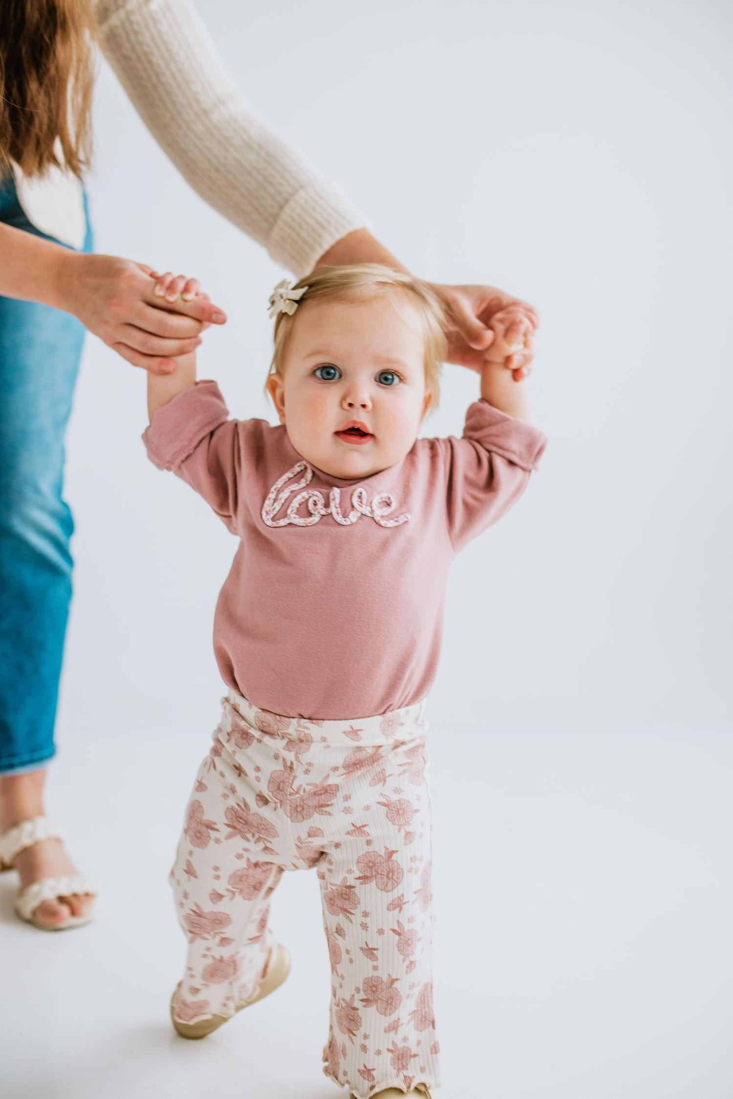 Adorable baby girl in pink love sweatshirt and floral pants taking first steps with mother's helping hands.